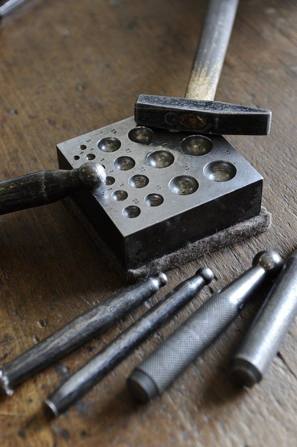 Jewelry-making tools: a steel block with circular depressions, hammers, and mandrels on a wooden surface.