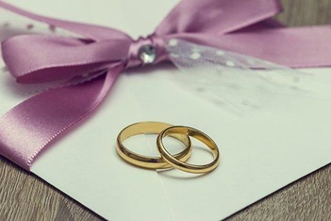 Two gold wedding rings on a white card, tied with a lavender ribbon and bow.