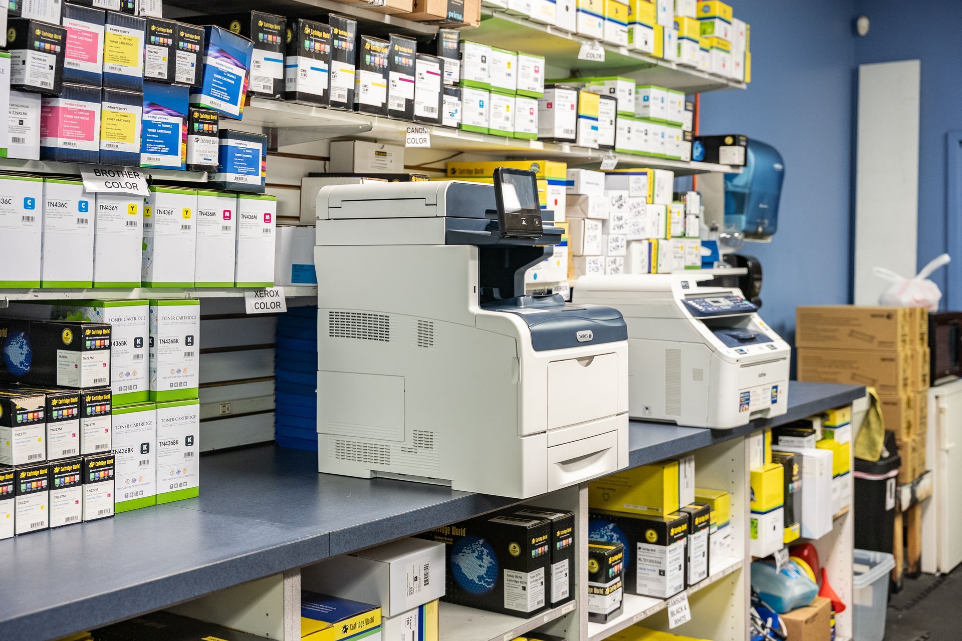 Printers and ink cartridges on a counter in a store with shelves of supplies.