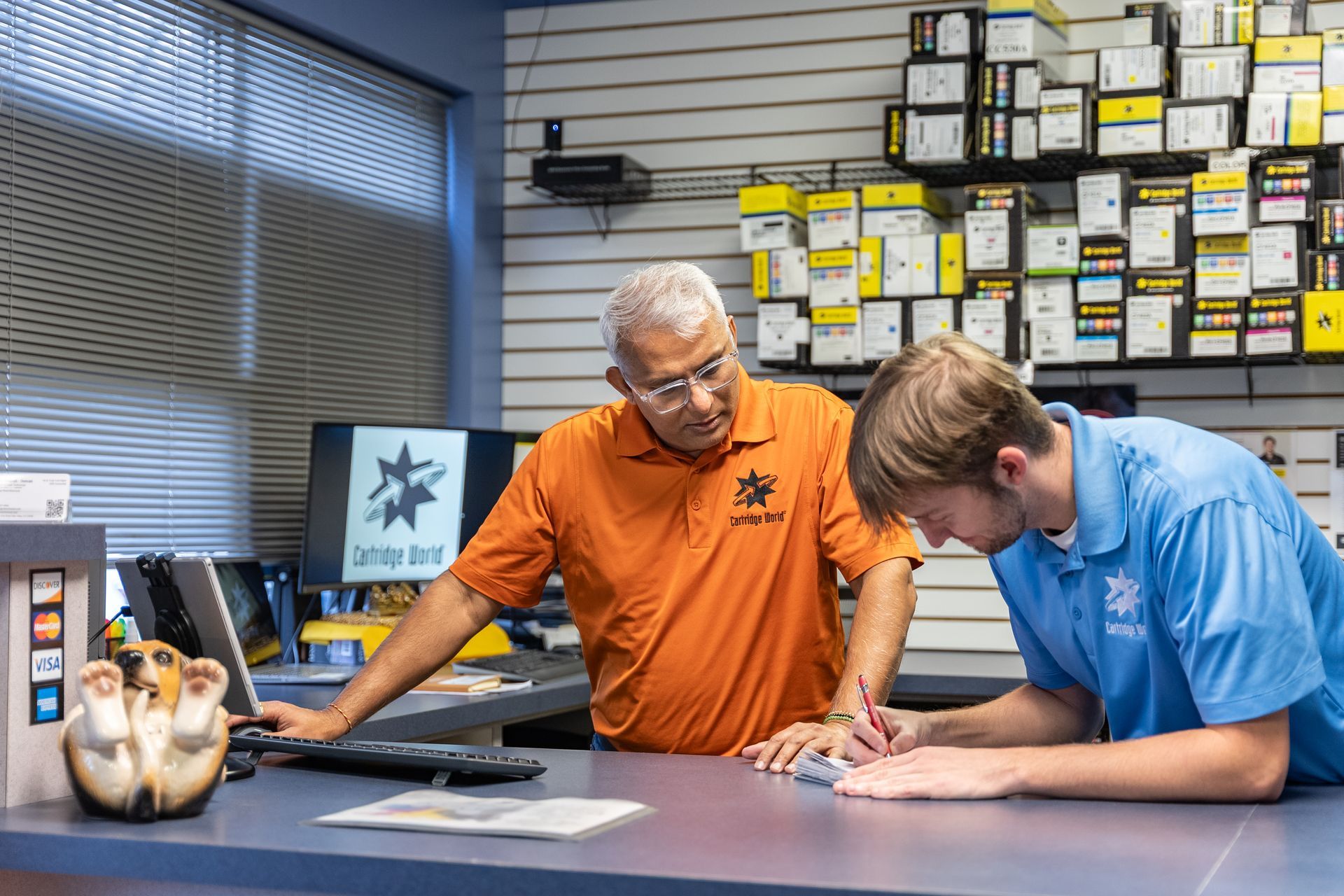 Two men at a counter in a store with parts; one man leans in to look at paperwork, the other looks on.