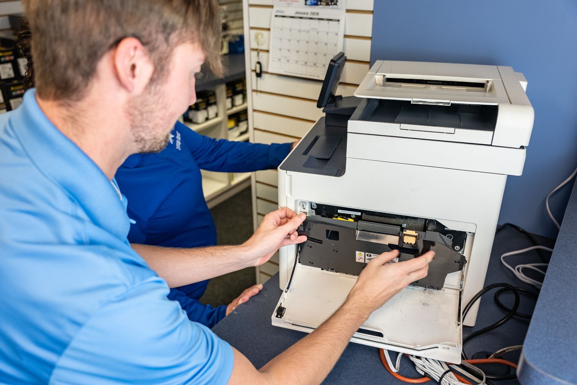 Man in blue shirt repairing a white printer, another person in the background.
