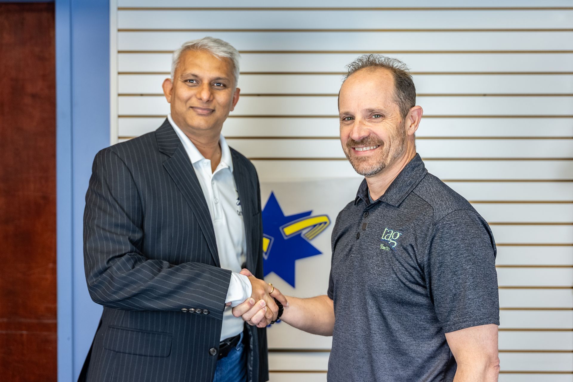 Two men shaking hands in front of a company logo. One in a suit, the other in a polo shirt.