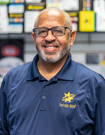 Man wearing glasses and navy shirt with a logo, smiling in a shop.