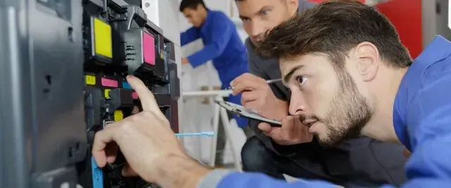Three technicians repairing a printer. One points at a cartridge. Others watch.