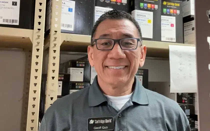Man with glasses smiles, standing in front of printer ink boxes. He wears a gray shirt with a nametag.