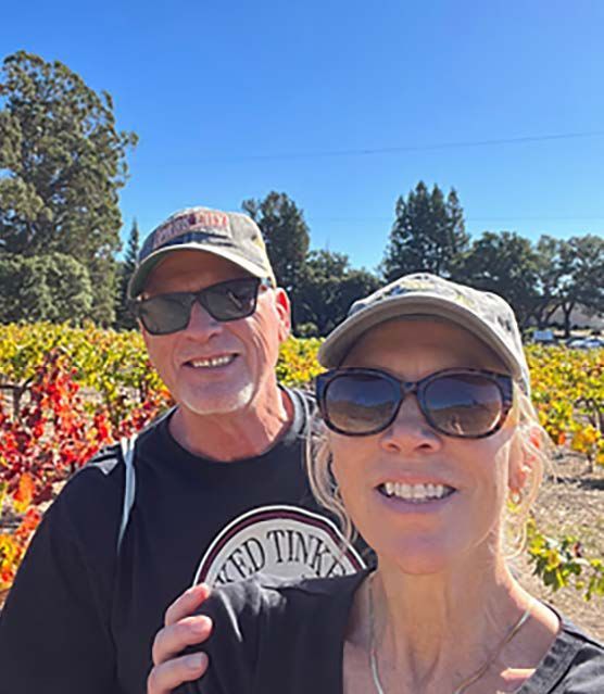 Couple smiling in vineyard, wearing hats and sunglasses. Fall colors in the background. Sunny day.