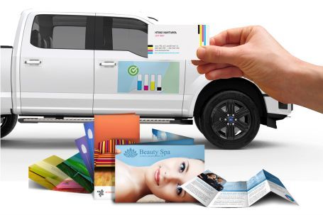 Hand holding a business card near a white truck with magnetic signs and other marketing materials scattered around.