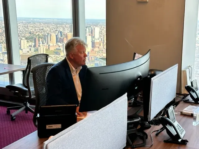 Man in suit at computer in high-rise office with city view, focused on monitor.