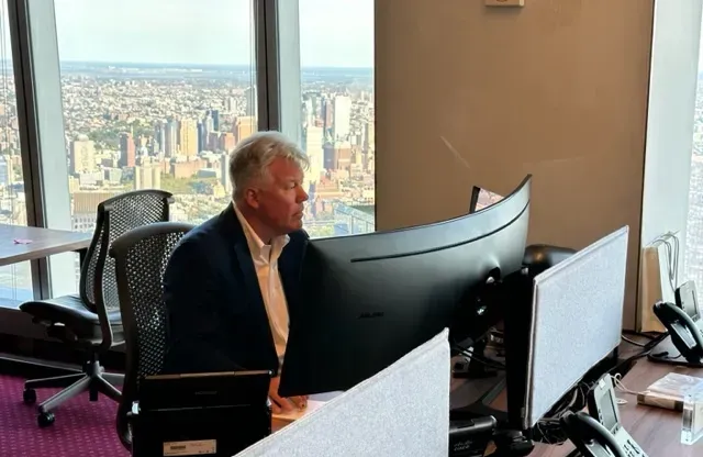 Man in suit jacket at computer in high-rise office overlooking city.