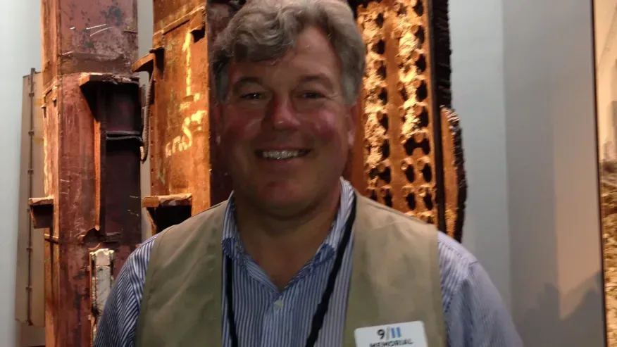 Man smiling, wearing a vest and badge, standing in front of structural steel fragments.