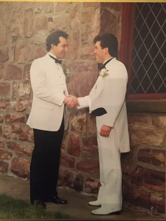 Two men in white tuxedos shake hands outside a stone building.