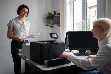Woman hands papers to colleague sitting at a desk with a printer; both smiling in office setting.
