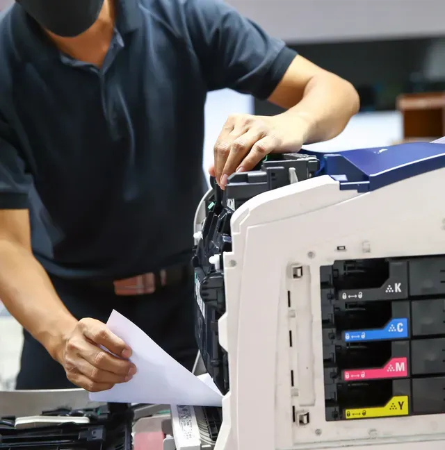 Person in a black mask fixing a printer, holding paper. Printer is white with color ink cartridges.