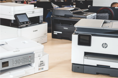 Printers of various brands on a desk: Canon, Brother, HP, and a smaller device. Office setting.