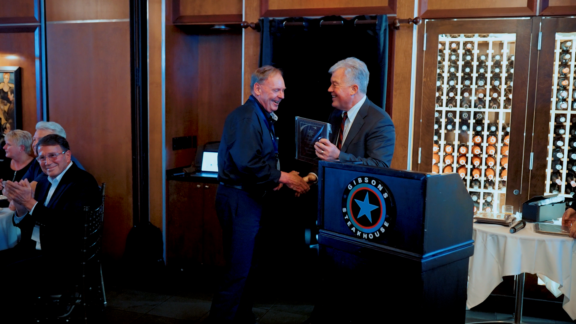 Man in blue shirt receives award from another at a podium, people applaud in a dining room.