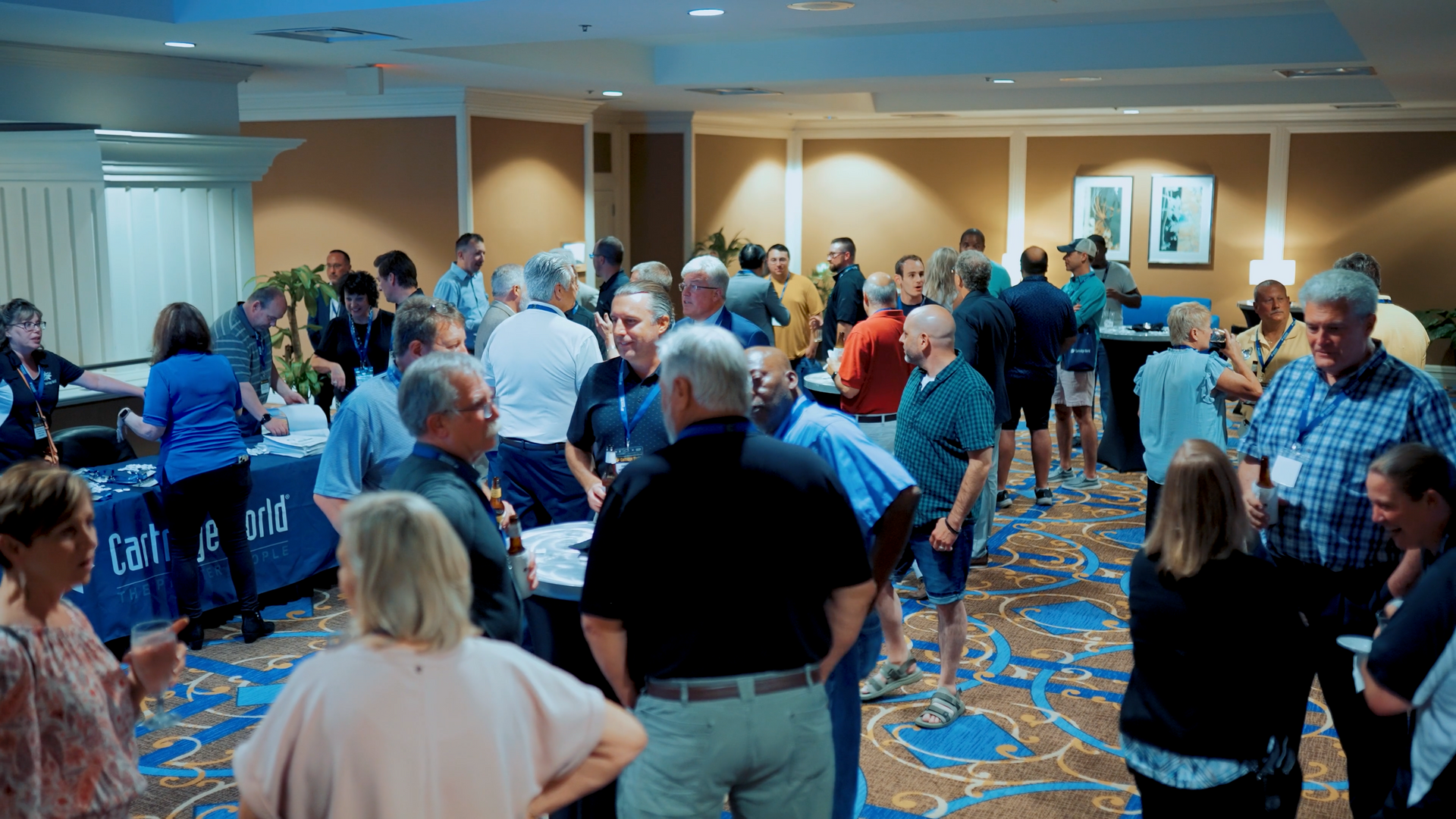 People at a conference mingle. Tables with informational materials, room with beige walls and patterned carpet.