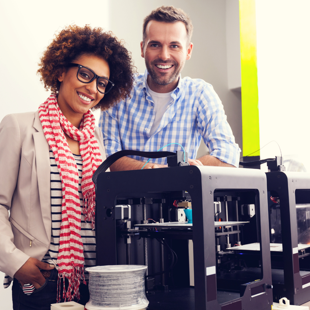 Woman and man smiling by a 3D printer. Woman wears glasses, striped shirt, scarf, blazer. Man wears plaid shirt.