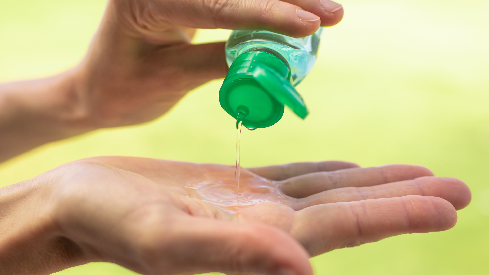 Hand pouring clear liquid sanitizer from a green-capped bottle into an open palm. Green background.