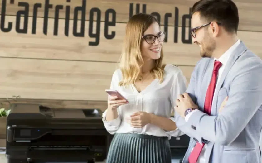 Woman holding phone and man in suit smiling, in front of a printer with the 