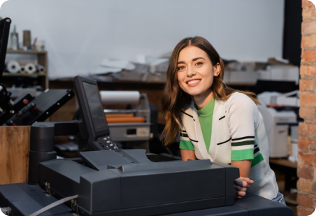 Woman smiles near printing equipment. She wears a white top with green accents and stands in a print shop.