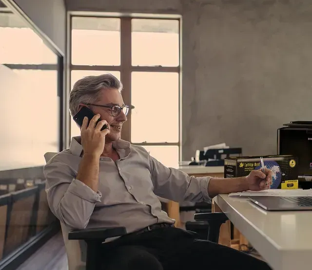 Man in glasses sits at desk, talking on phone, smiling. Office setting.