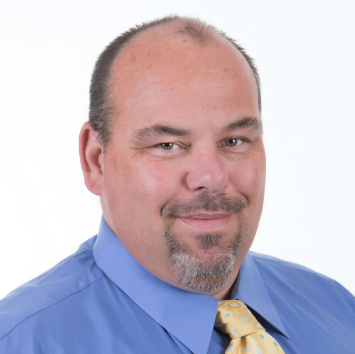 Man in a blue shirt and yellow tie smiles at the camera, against a white background.