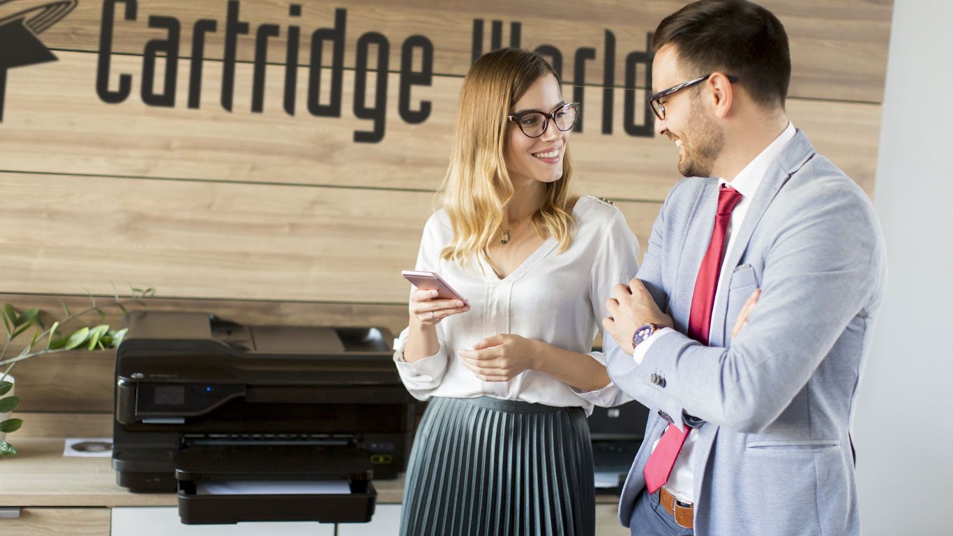 Woman and man looking at each other, in an office. They both wear glasses and smile near a printer at Cartridge World.