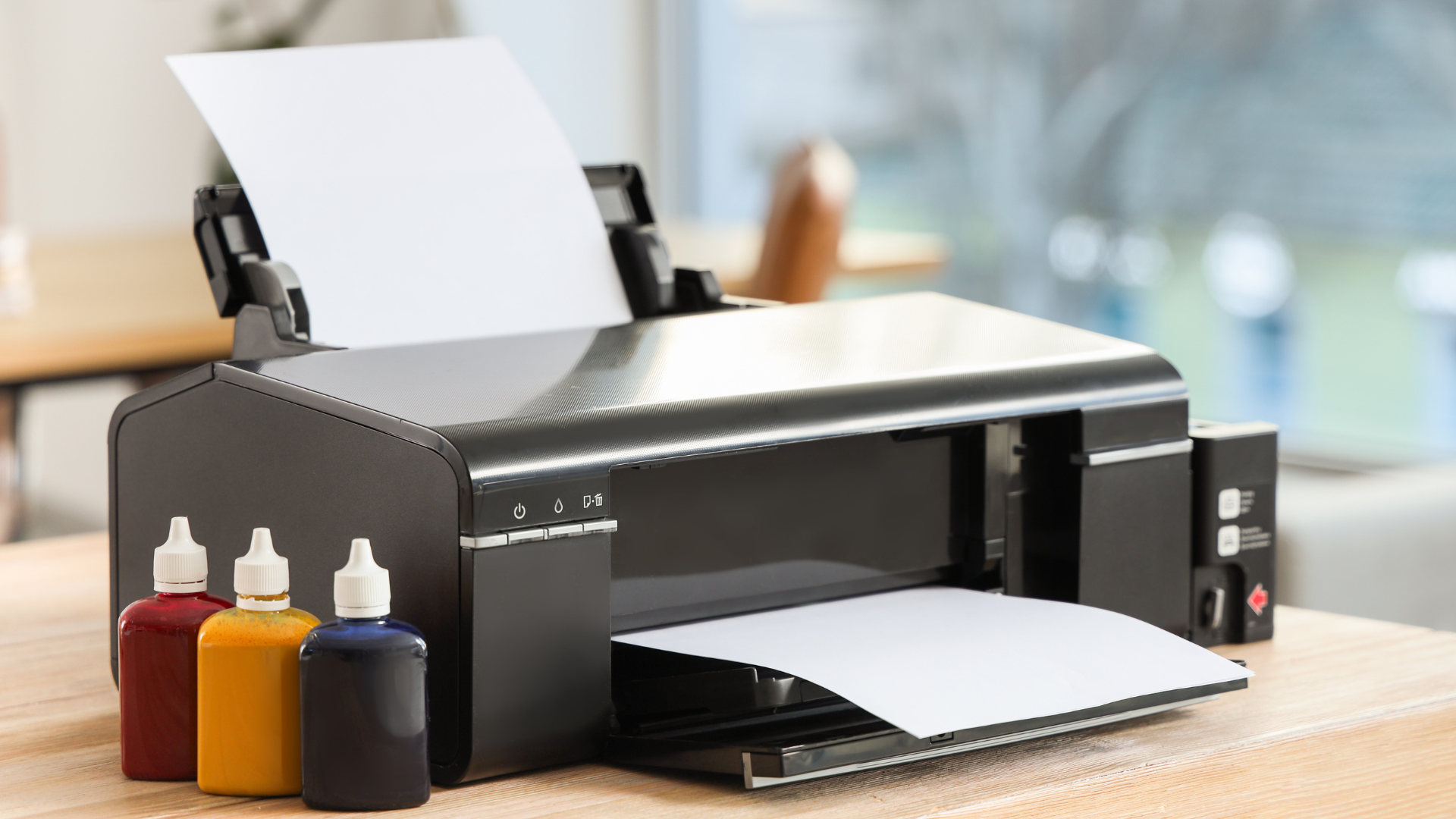 Black inkjet printer with open paper tray, next to three ink bottles: red, yellow, and blue.