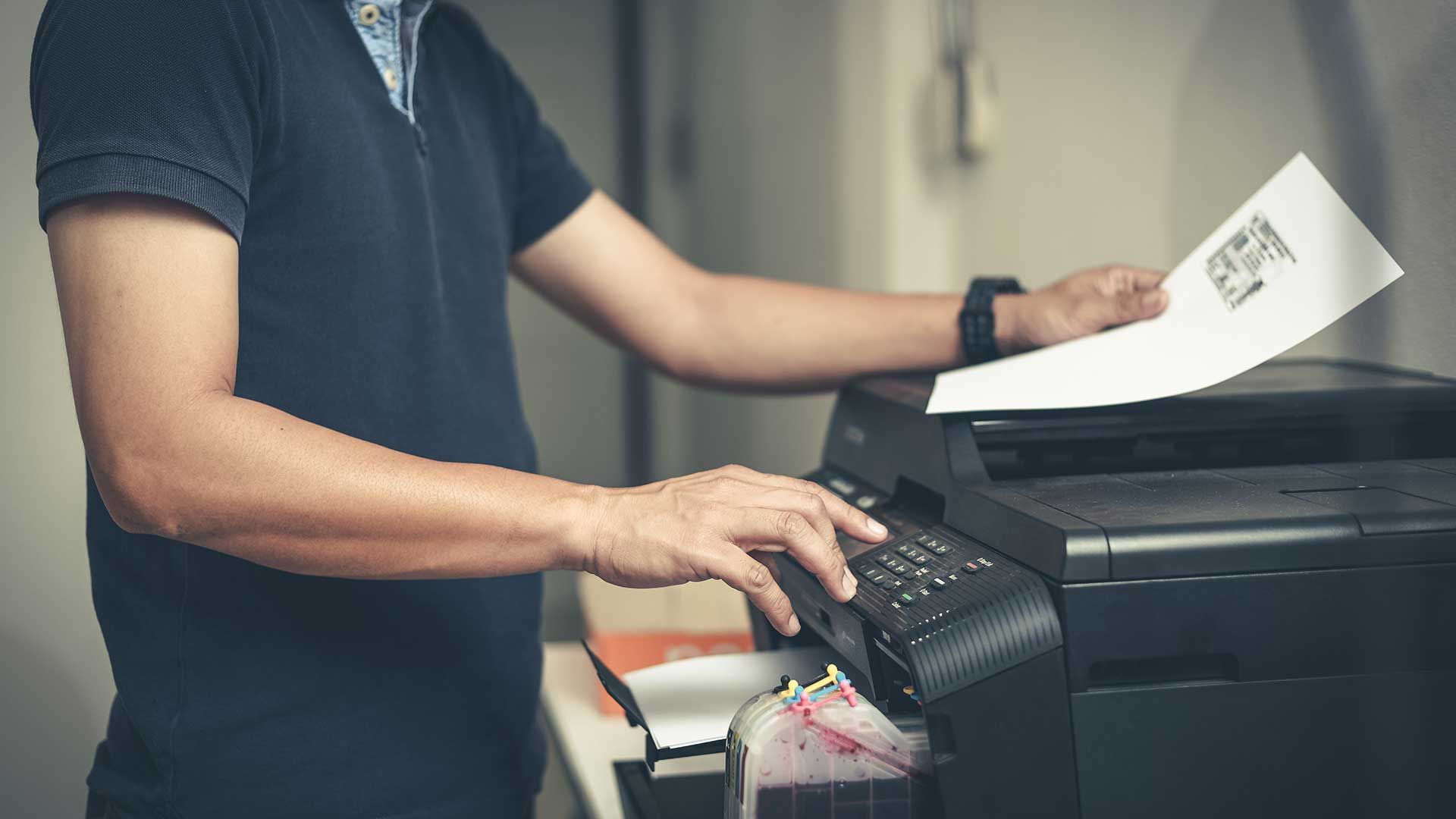 Person operating a black printer, holding a printed document indoors.