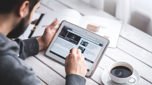 Person using a tablet on a white wooden table next to a cup of coffee.