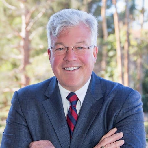 Man in a suit smiles, arms crossed, in a sunny outdoor setting with trees.