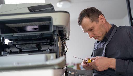 Man repairs a printer with a screwdriver in a brightly lit office setting.
