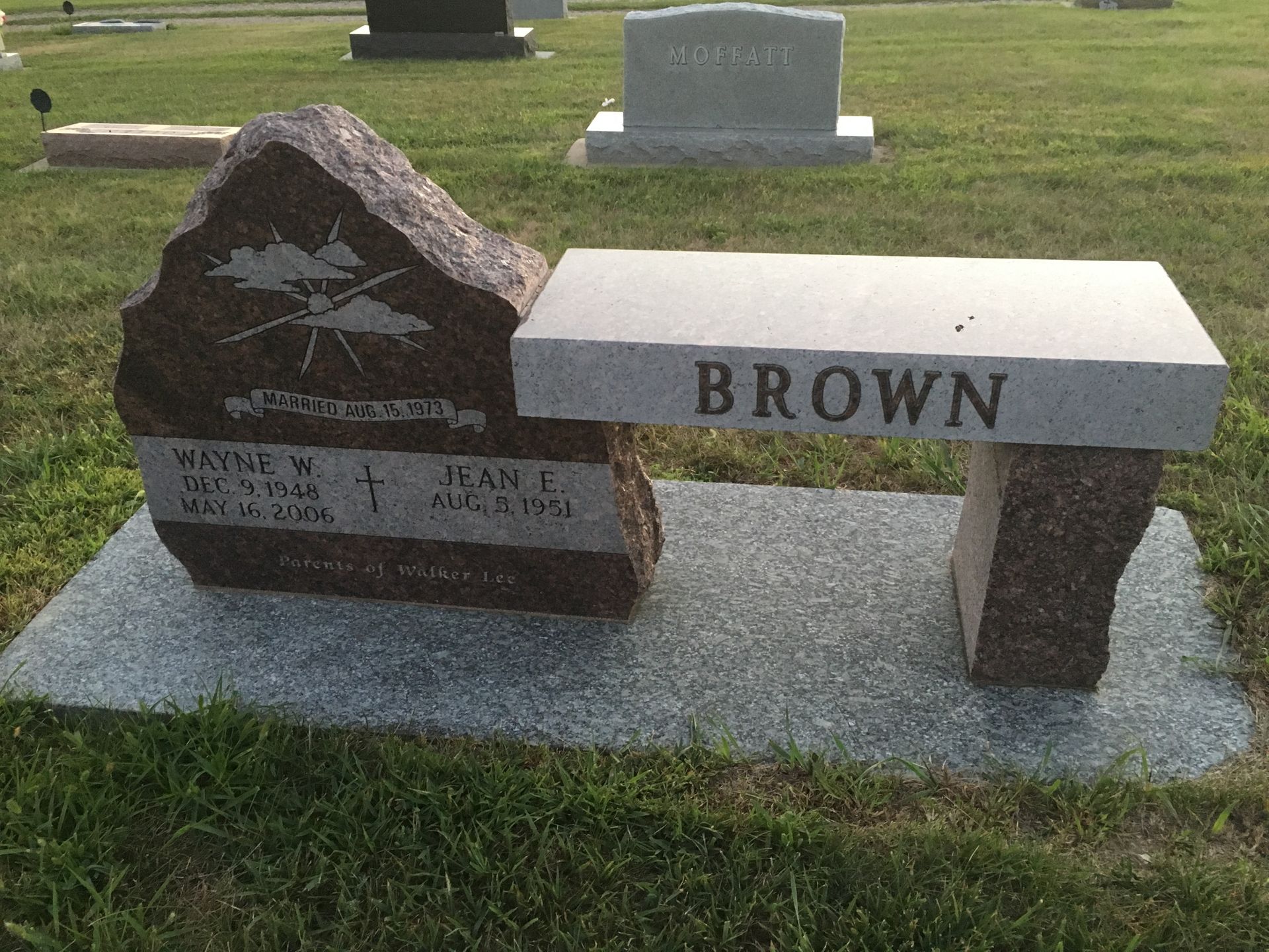 Brown memorial bench and stone grave marker in a cemetery lawn