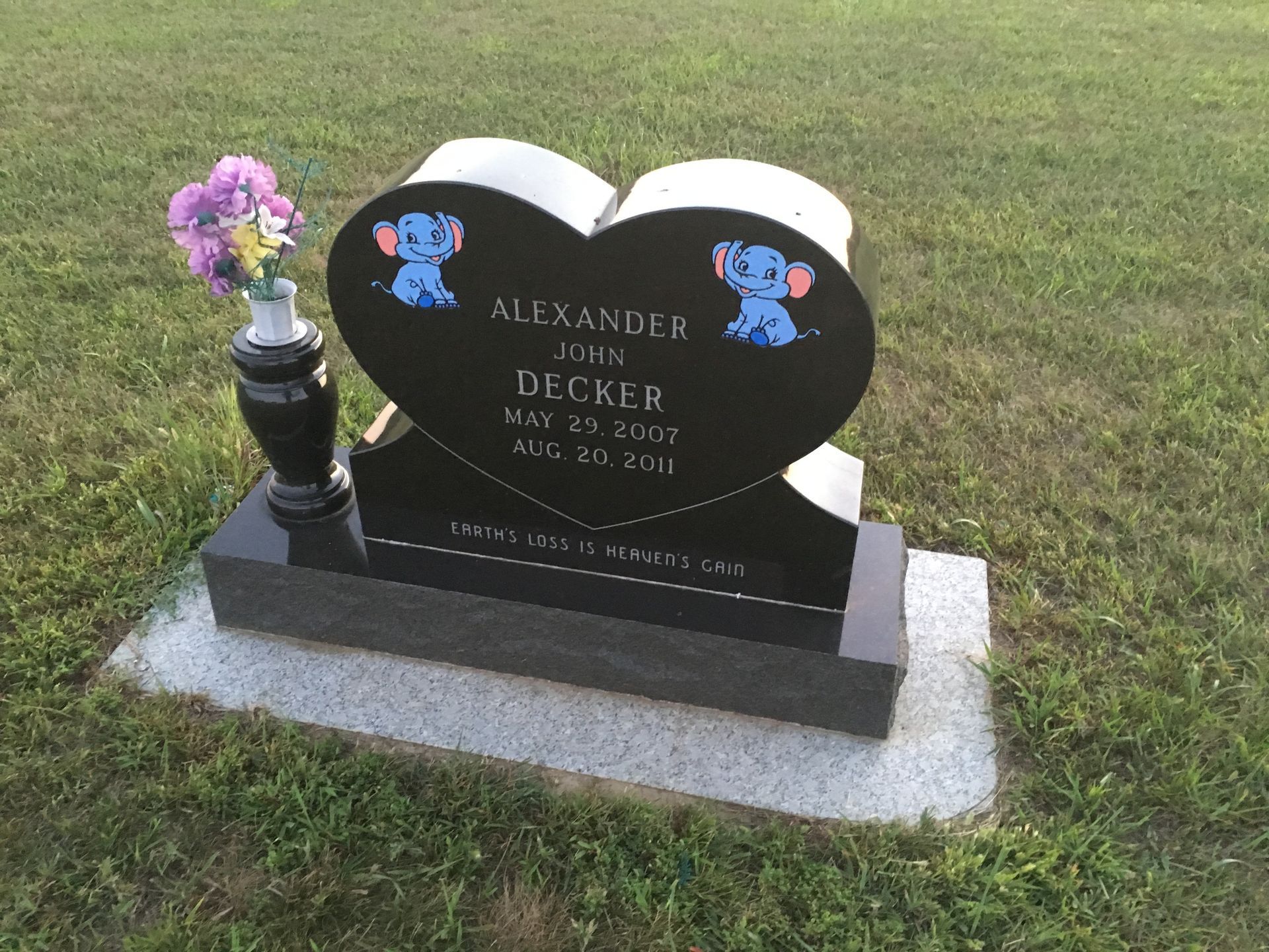 Heart-shaped black gravestone with blue teddy bears and flowers in a grassy cemetery.