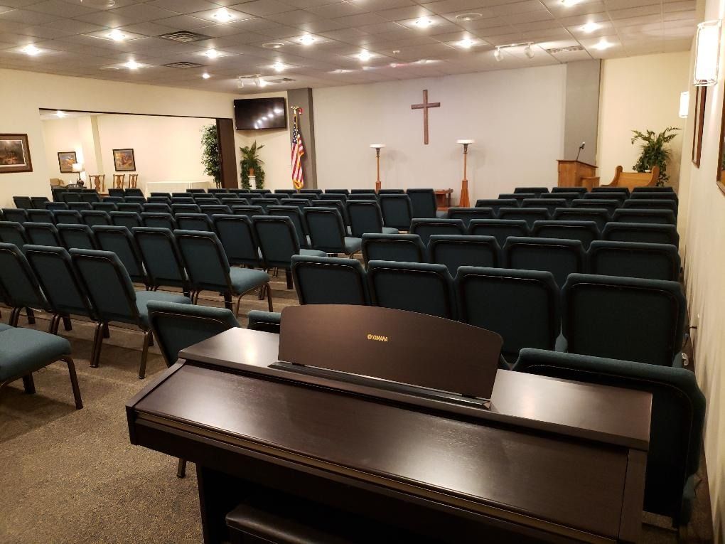 Empty church sanctuary with rows of chairs, a piano in front, and a cross on the wall.