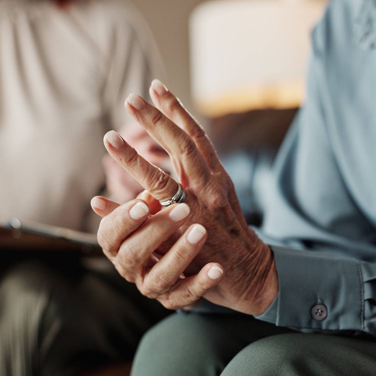 Person's hands with a ring, resting on a lap; another person sits in the background.