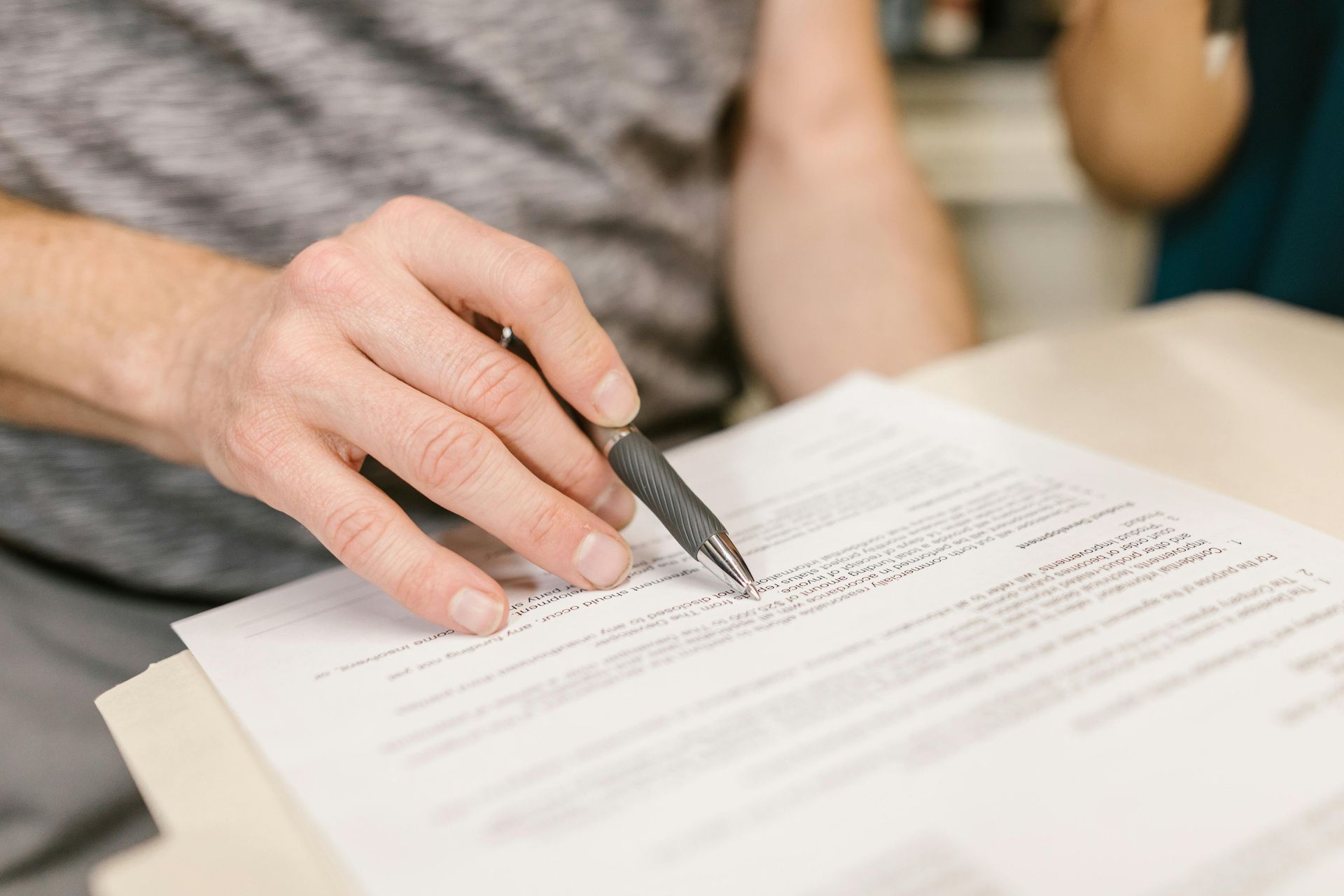 Person's hand pointing pen at document, possibly reading or reviewing, with another person in background.