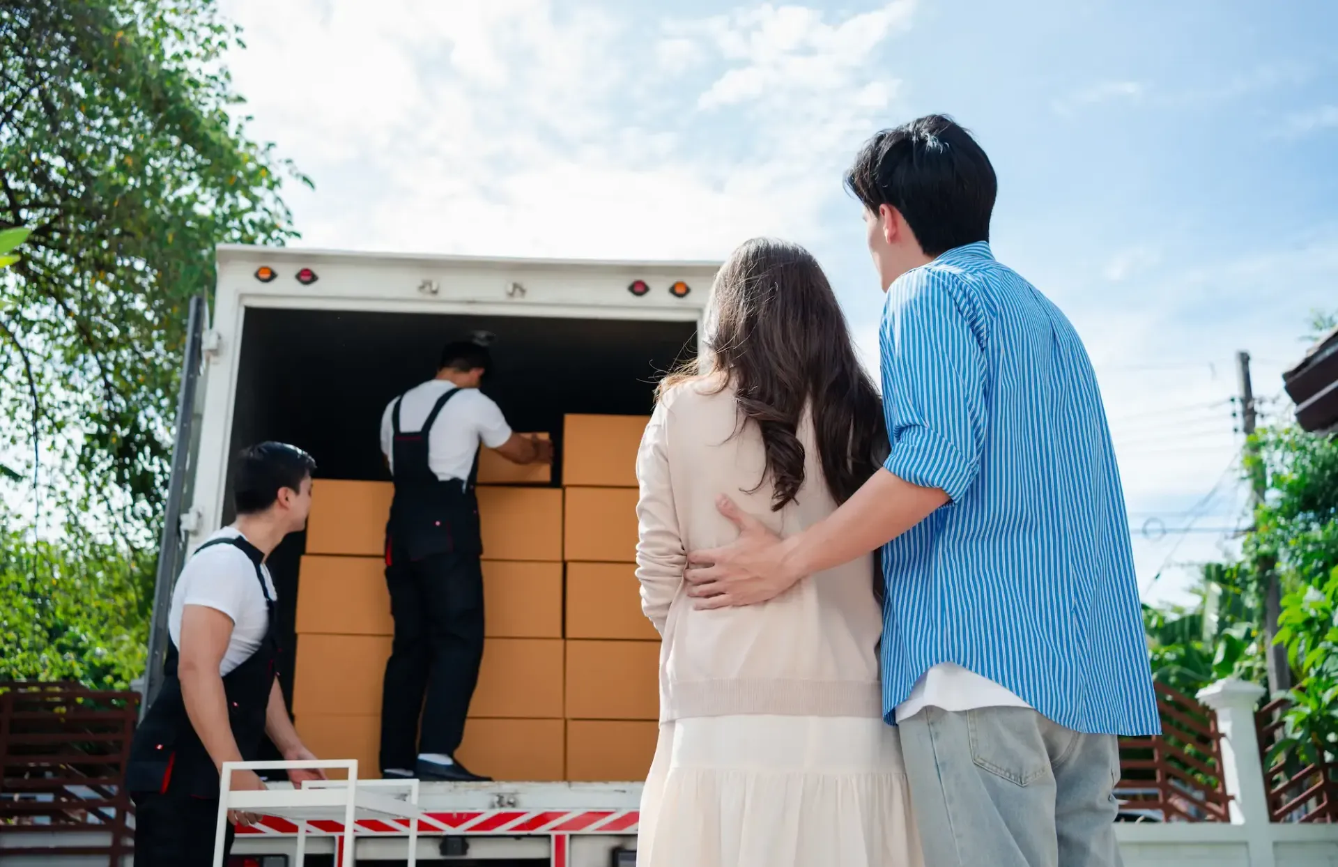 Couple watches movers load boxes into a truck. Sunny day, blue sky.