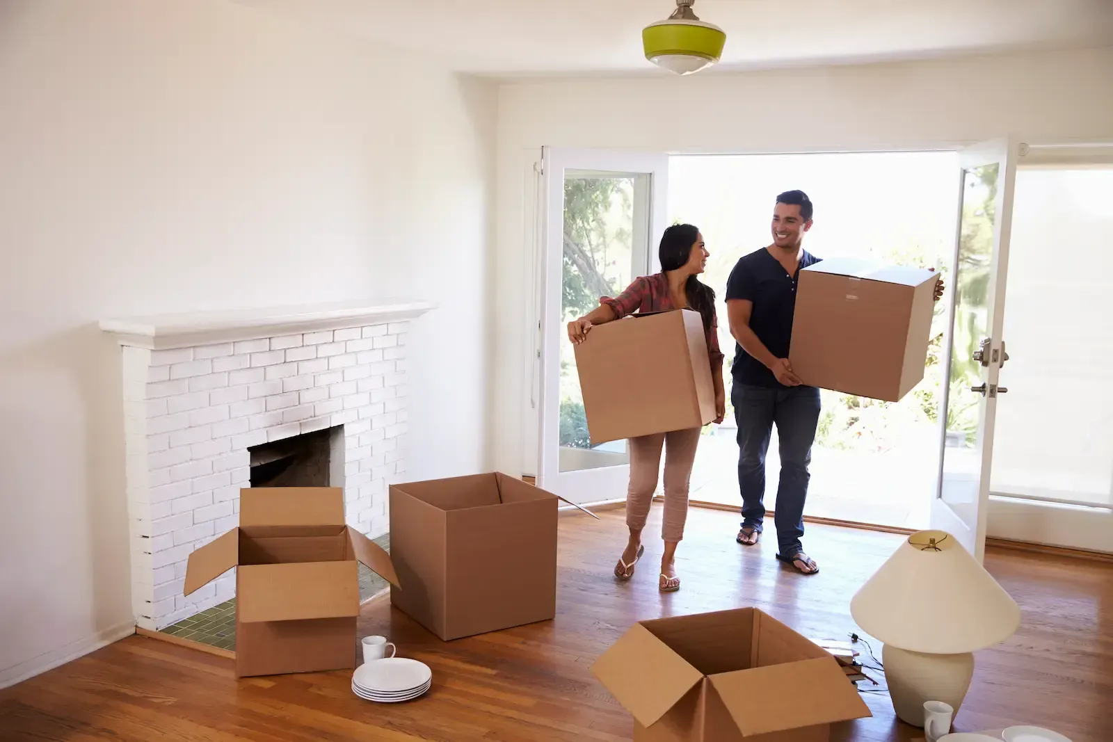 Two people carrying boxes in a bright, empty room with a fireplace and moving supplies.