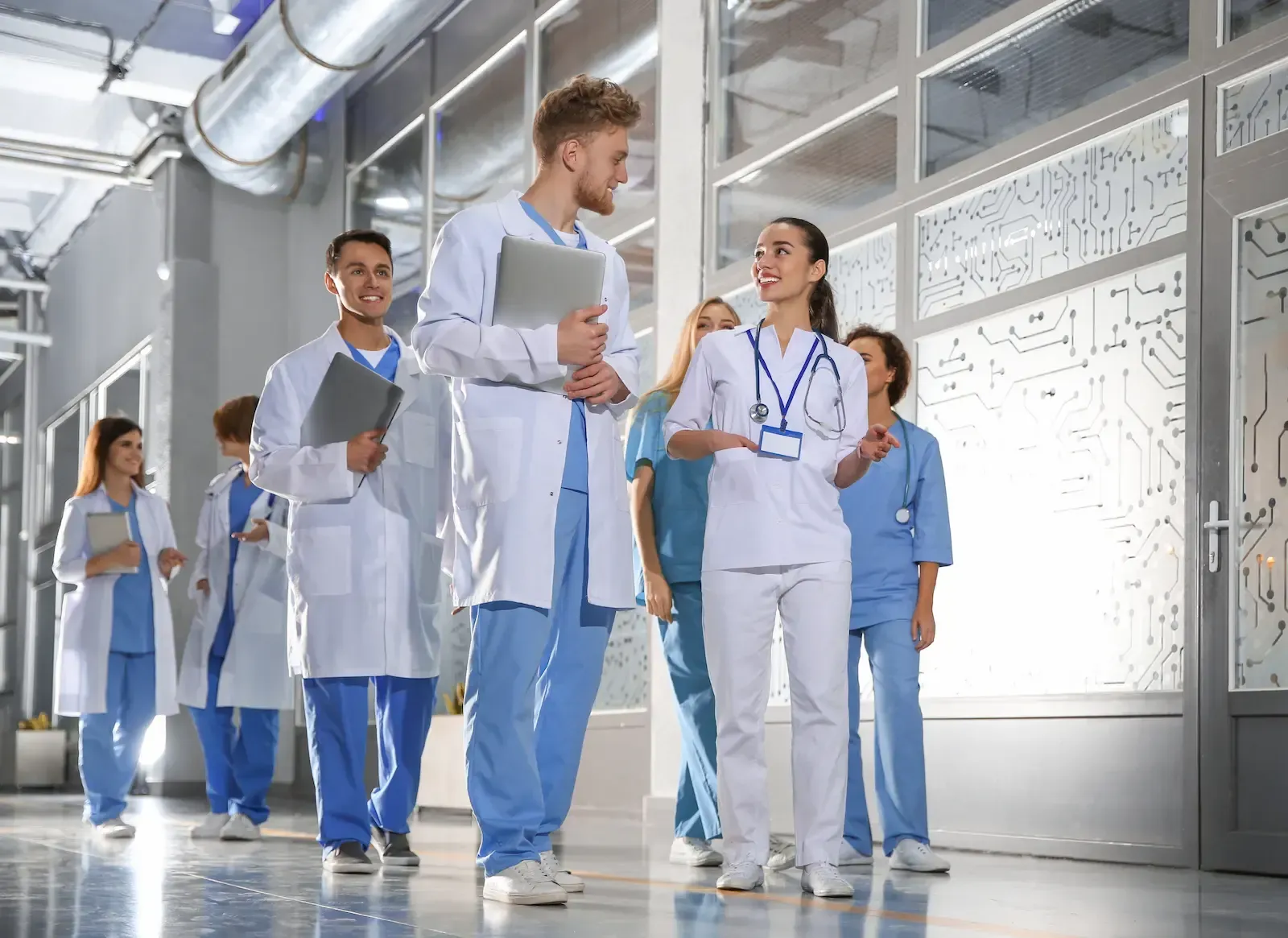 Healthcare workers in white and blue scrubs walking and talking in a bright hospital corridor