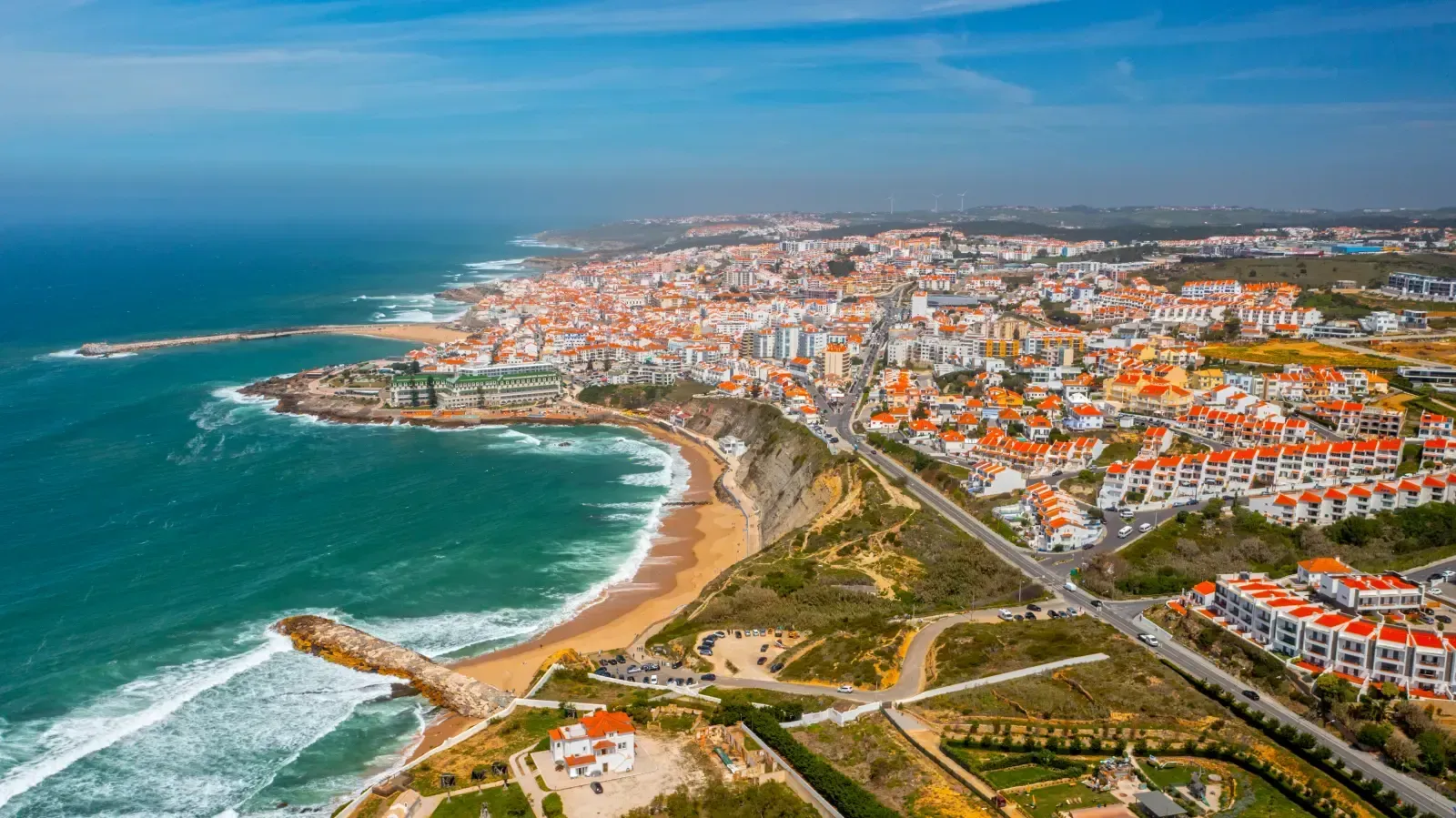 Aerial view of a coastal town in Portugal featuring terracotta rooftops, a sandy beach, and waves hitting rocky shores.