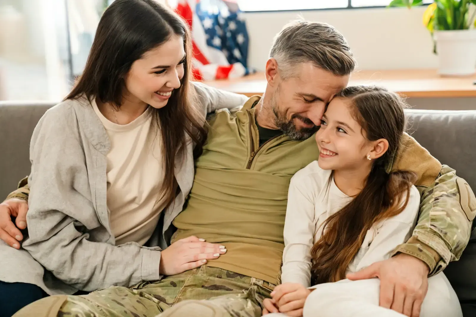 A soldier in uniform sits on a sofa, smiling and embracing a woman and a child. They are together in a bright room.