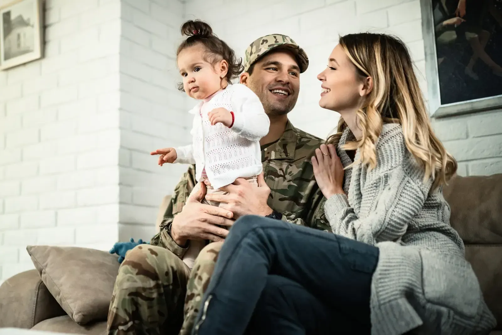 Man in uniform holds child as woman smiles beside them on a couch.