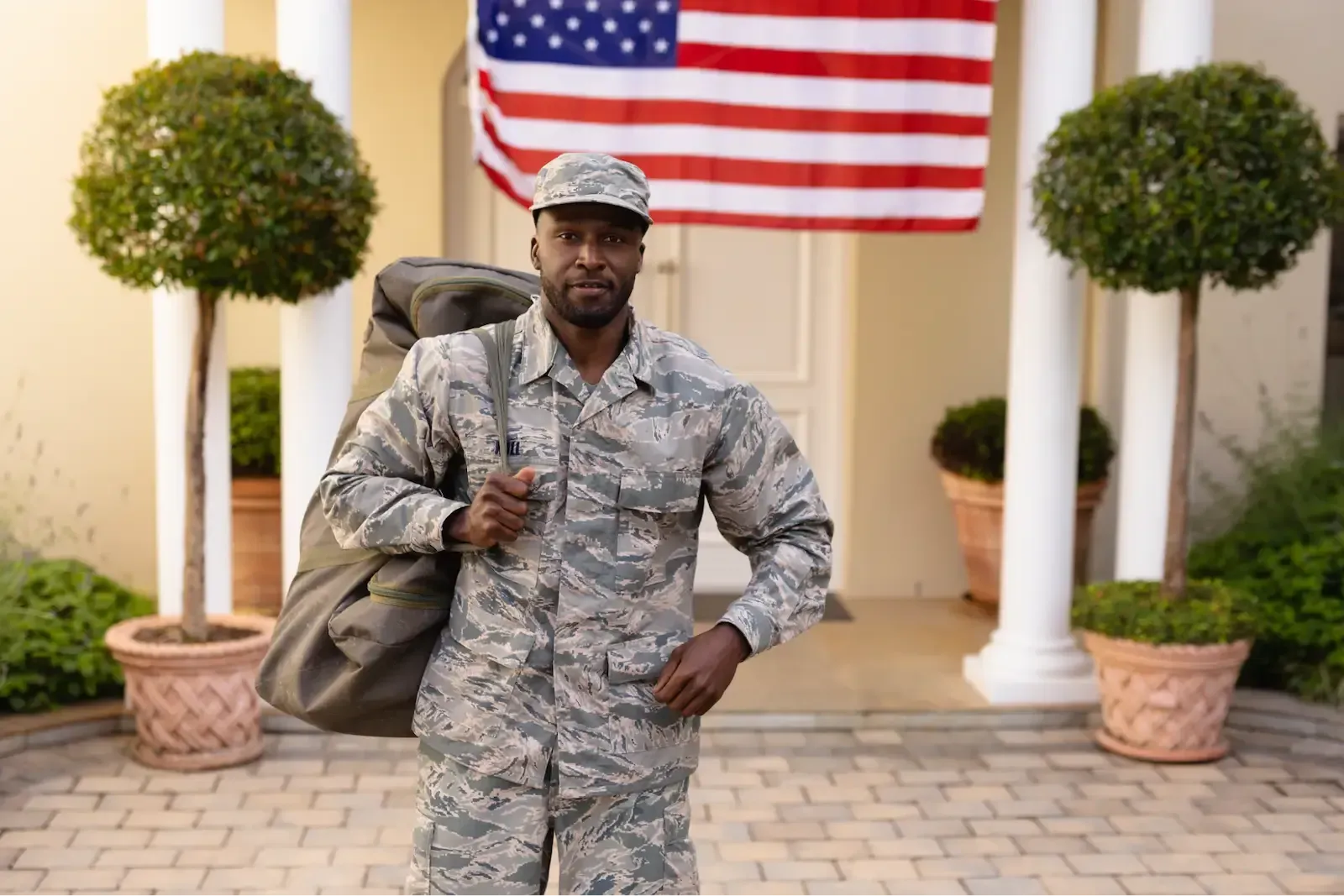 A person in military uniform stands outside a house under an American flag, carrying a bag.
