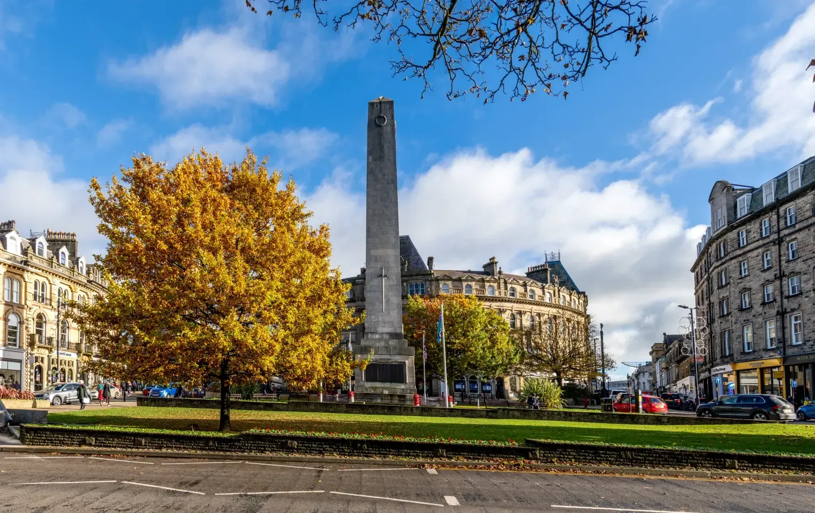 A tall stone obelisk war memorial stands in a park square with a vibrant yellow autumn tree against a bright blue sky.