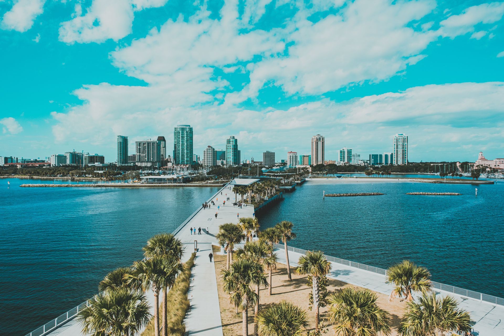 Palm-lined causeway over blue water leading to a city skyline under a bright sky