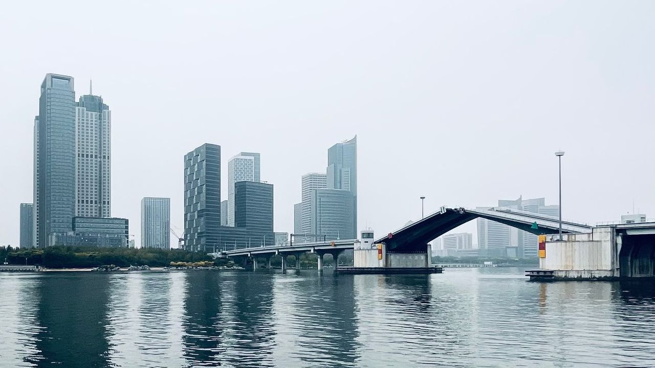 City skyline with modern towers reflected on calm water beside a raised bridge.