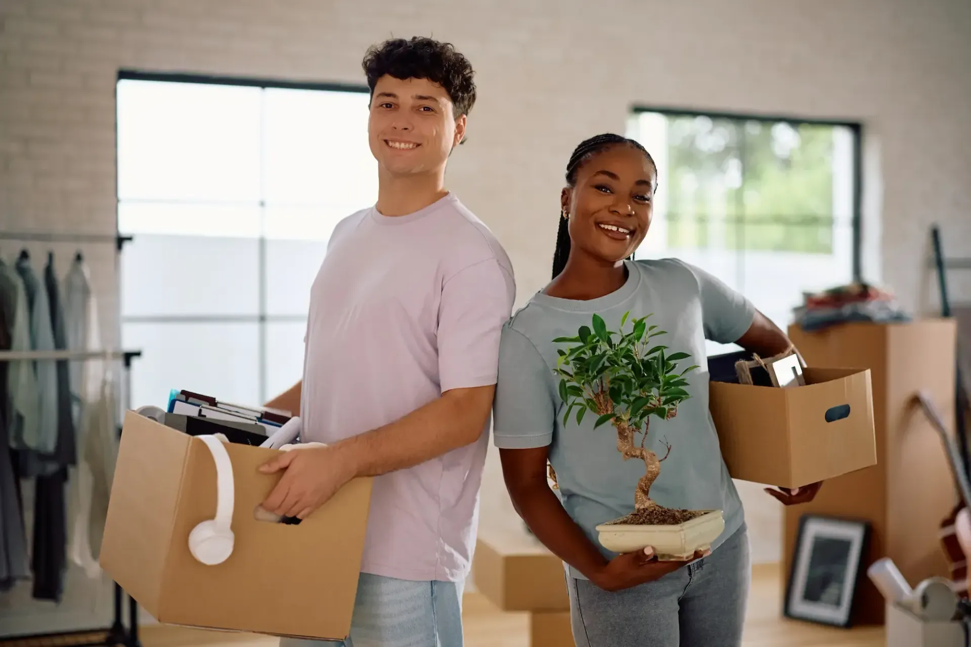 Couple holding boxes, smiling, moving into a new home. One holds a small tree, the other headphones.