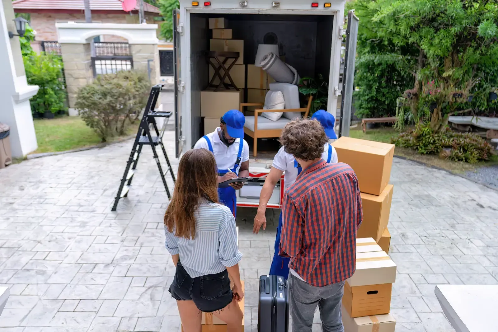 Movers loading a truck with boxes and furniture; two people watch on a paved driveway.