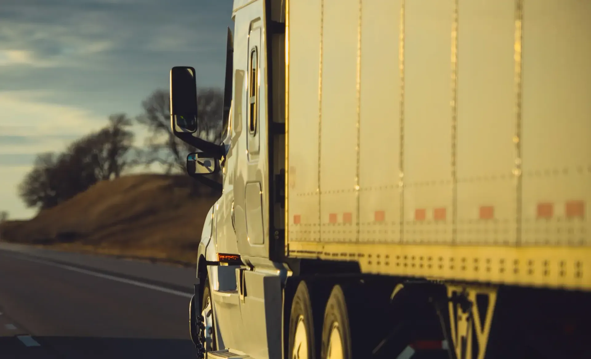 Semi-truck driving on a highway, side view. White cab, yellow trailer, and a blurred landscape.